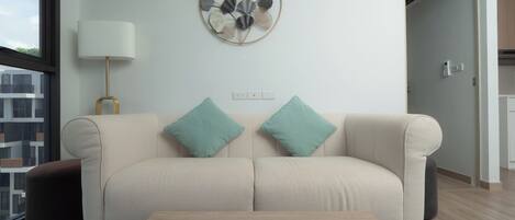 Un salón moderno con un sofá color beige, cojines turquesa, una mesa de centro de madera con un jarrón azul, un aire acondicionado instalado en la pared y una obra de arte con hojas decorativas.
