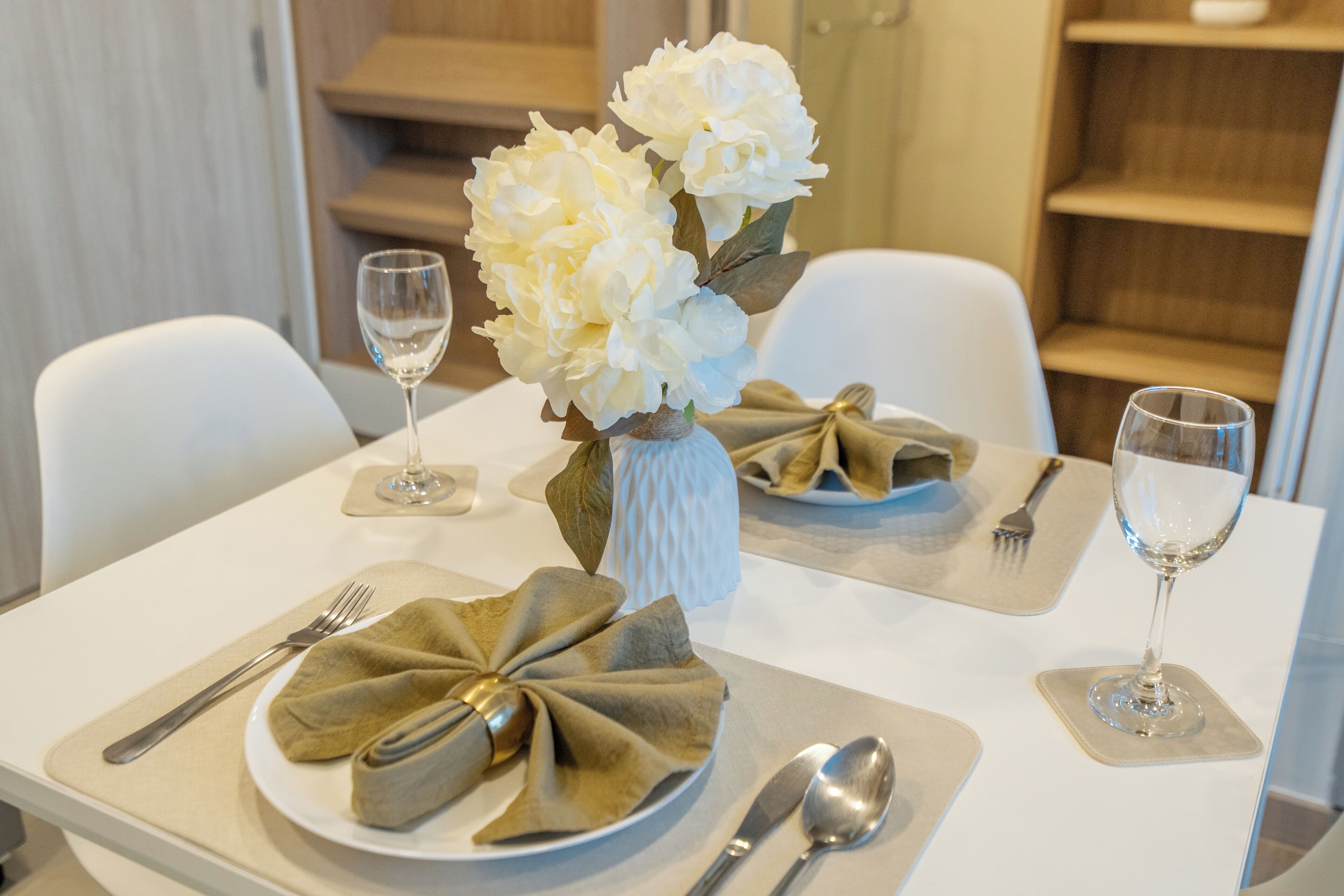 A dining table set with white plates, folded napkins, cutlery, and a vase with white flowers.