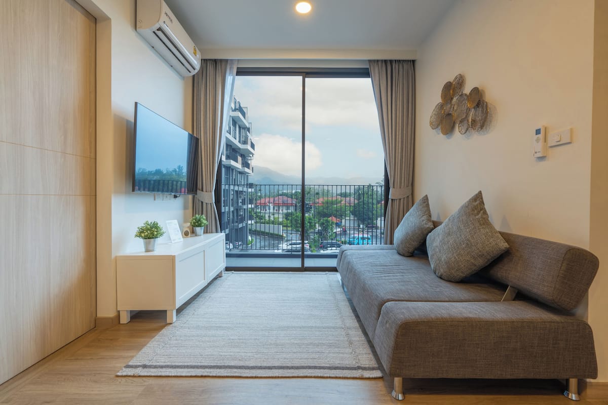 Una sala moderna con sofá, televisor de pantalla plana, una mesita auxiliar y vista a un edificio a través de un ventanal grande.
