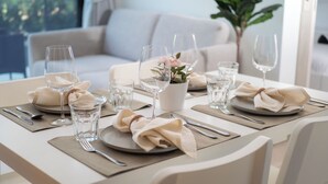 A dining table set with plates, glasses, and cutlery, surrounded by white chairs in a well-lit room with a sofa and a plant in the background. - Laguna SkyPark new brand apartments (Choeng Thale)