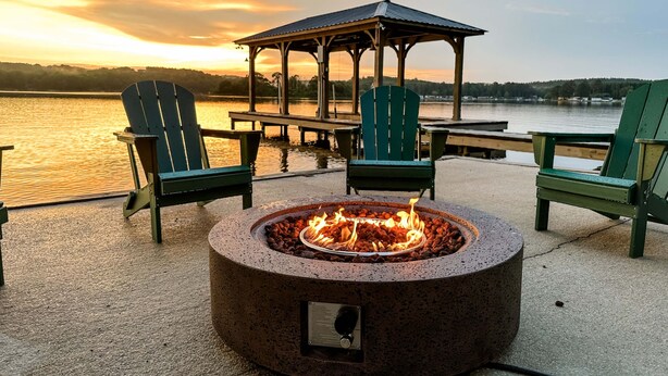 A circular fire pit with a metal cover, surrounded by green Adirondack chairs, a wooden gazebo, and a lakeside view. - Mallards Nest on Lay Lake: Game Room & Boat Ramp (Shelby)
