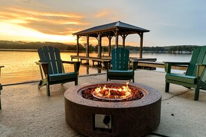 A circular fire pit with a metal cover, surrounded by green Adirondack chairs, a wooden gazebo, and a lakeside view.