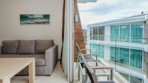 Una sala de estar moderna con un sofá gris, una mesa de centro de madera y una barandilla de cristal con vistas a un edificio de grandes ventanales.