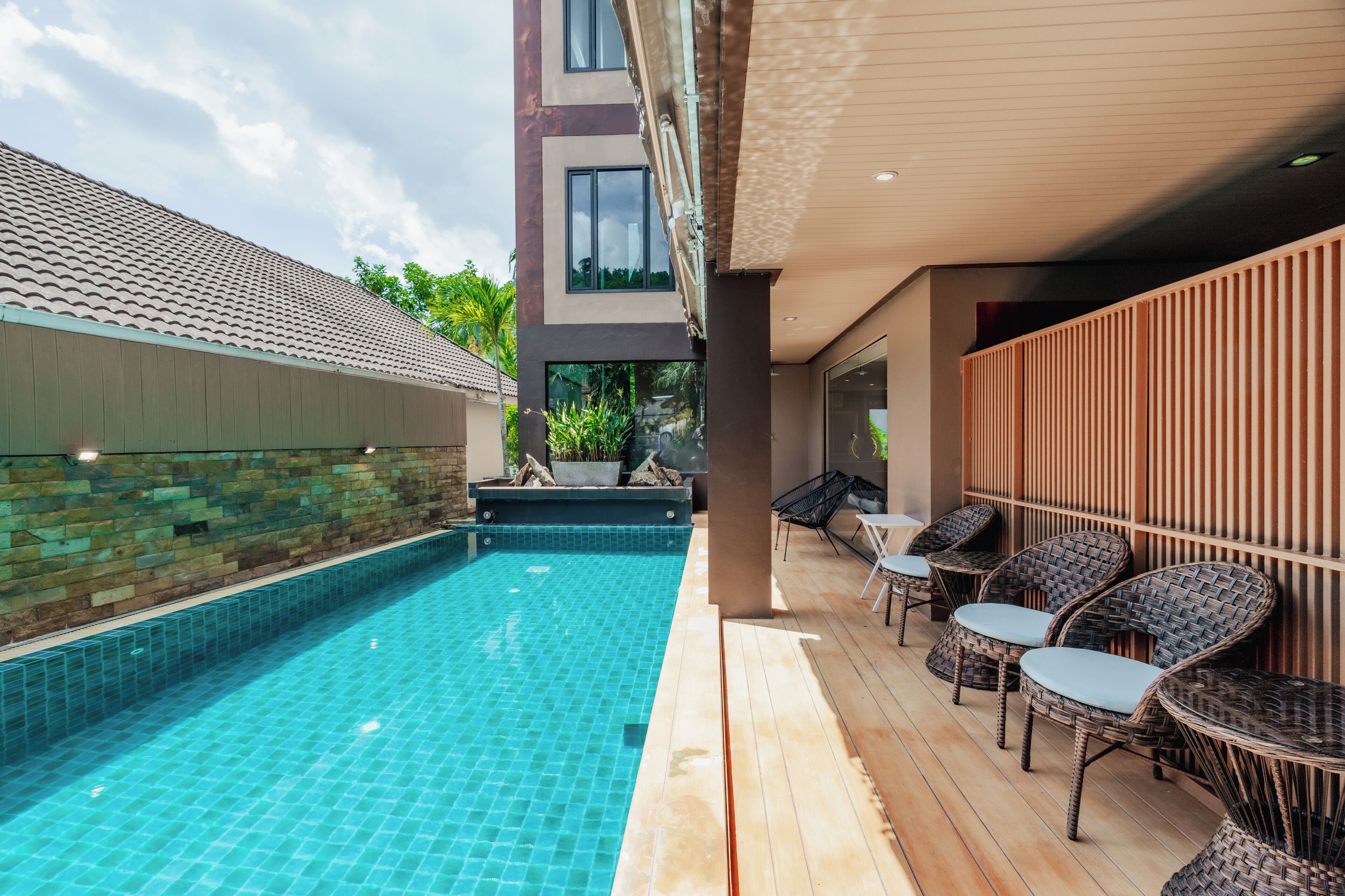 Une piscine avec une terrasse carrelée, des fauteuils en osier et un bâtiment moderne doté de grandes fenêtres.