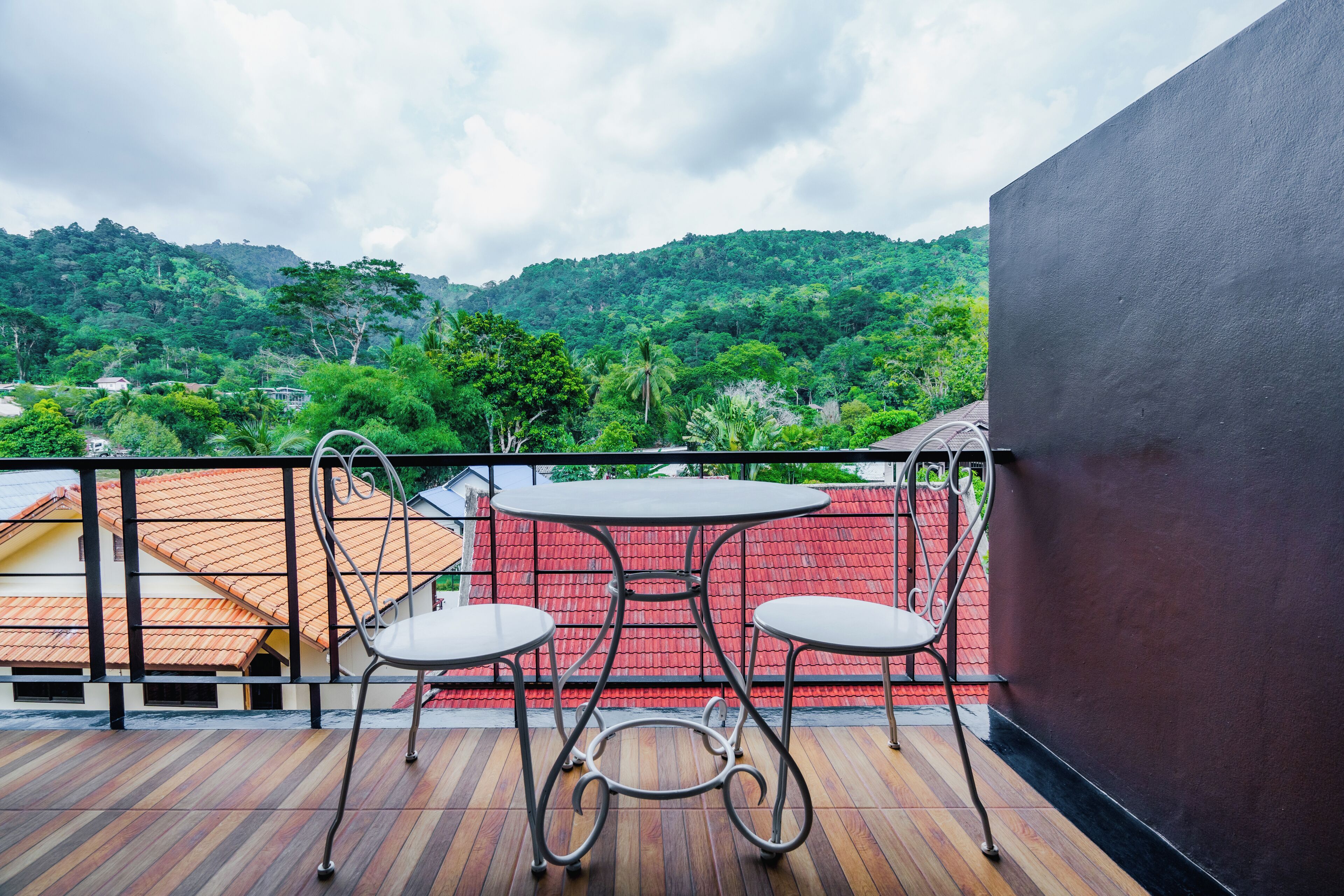 Un balcón con mesa y sillas, con vistas a un paisaje verde exuberante y un edificio con tejados rojos.