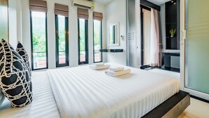 A modern hotel room with a large bed, a seating area with a woven chair, and a view of greenery through the windows. - Westkey Kamala Hotel (Kamala)