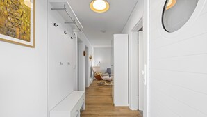 Un pasillo moderno con paredes blancas, suelo de madera y un banco. Hay luces de techo y un espejo redondo a la derecha.