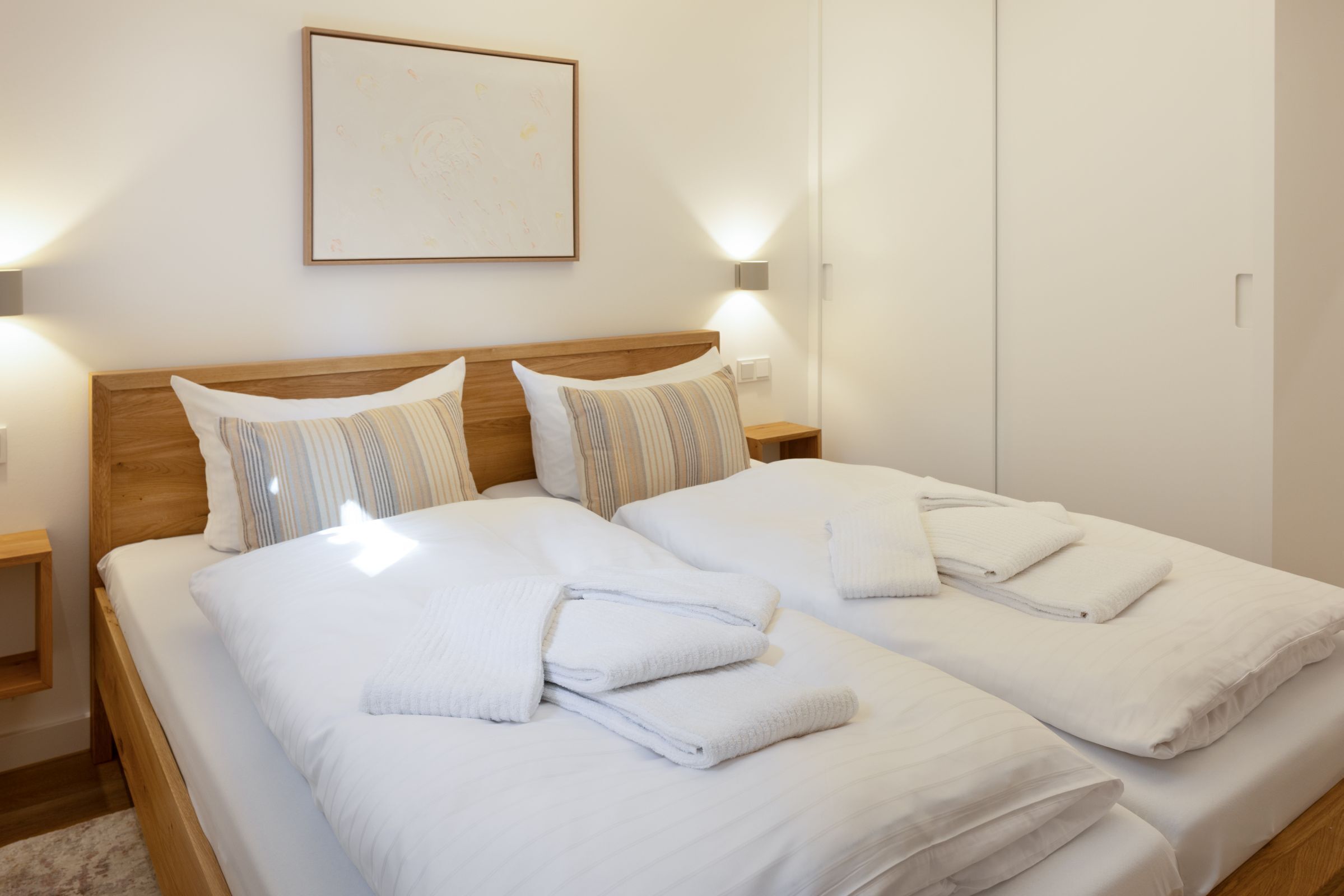 A double bed with white linens and striped pillows, a wooden headboard, and two bedside tables with wall-mounted lamps.