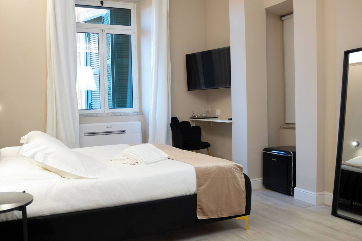 Un dormitorio con cama, mesita de noche, ventana, aire acondicionado, televisor y espejo.