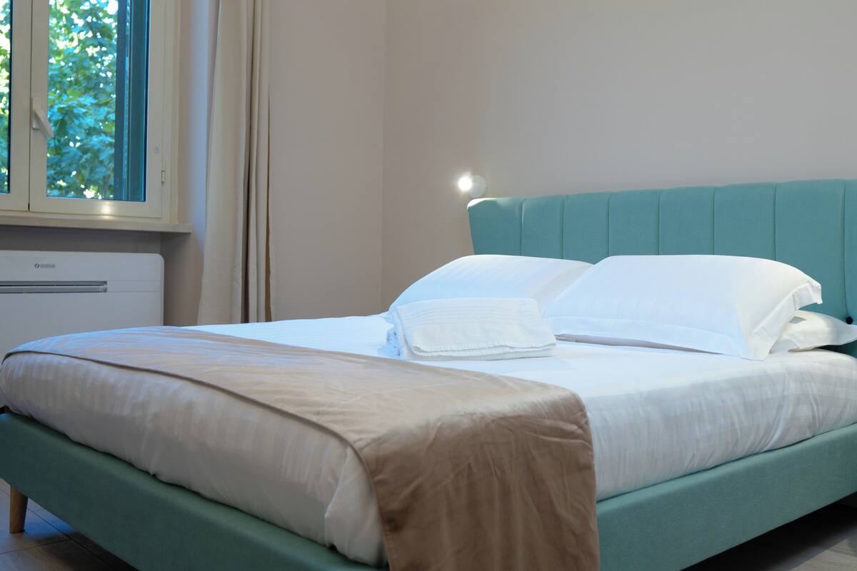 Una cama bien tendida con sábanas blancas y una manta color beige, un cabecero verde azulado y una mesita de noche con una lámpara.