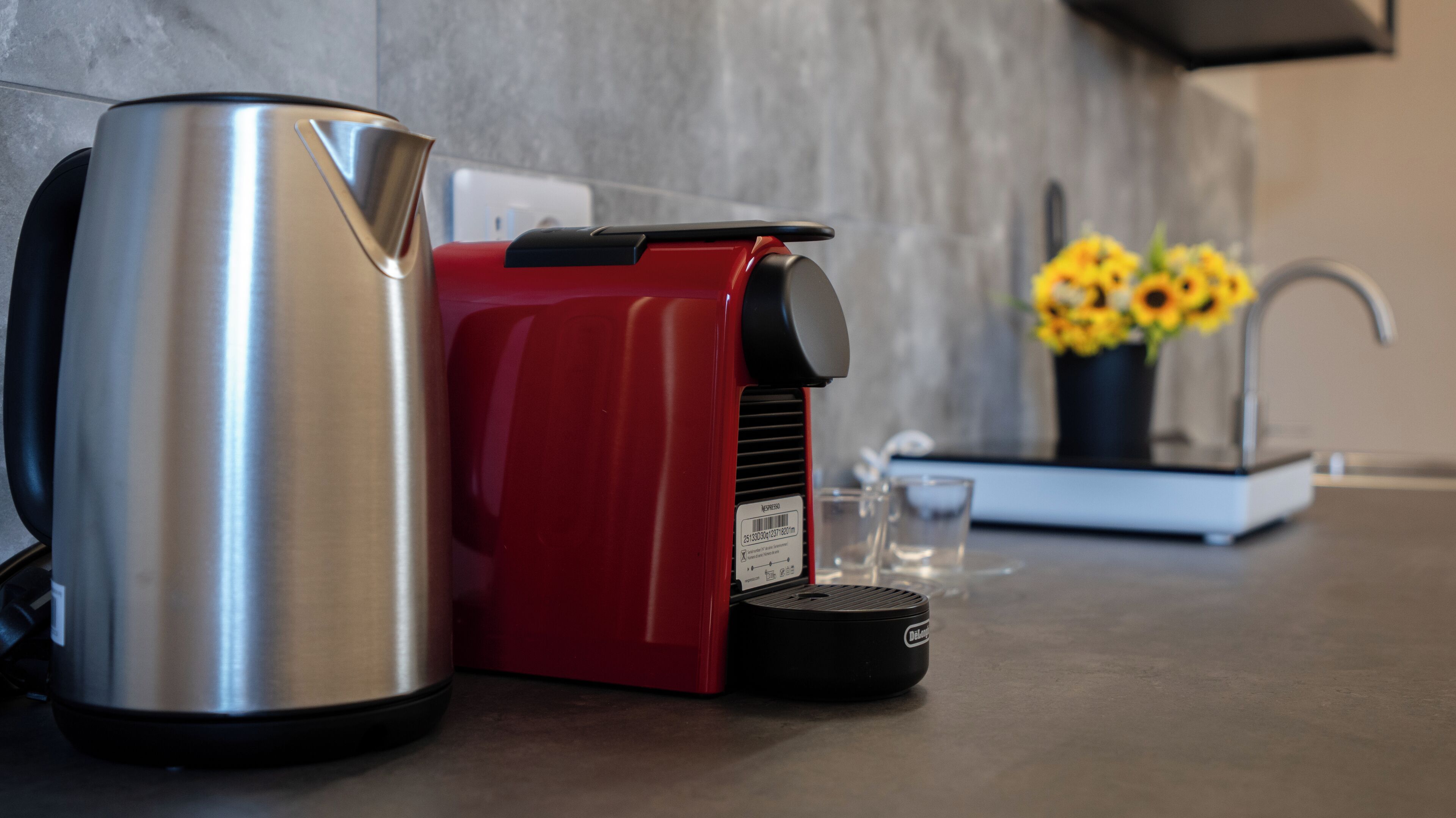 Una cocina con un hervidor de acero inoxidable, una cafetera roja, un vaso y un jarrón con flores amarillas sobre la encimera.