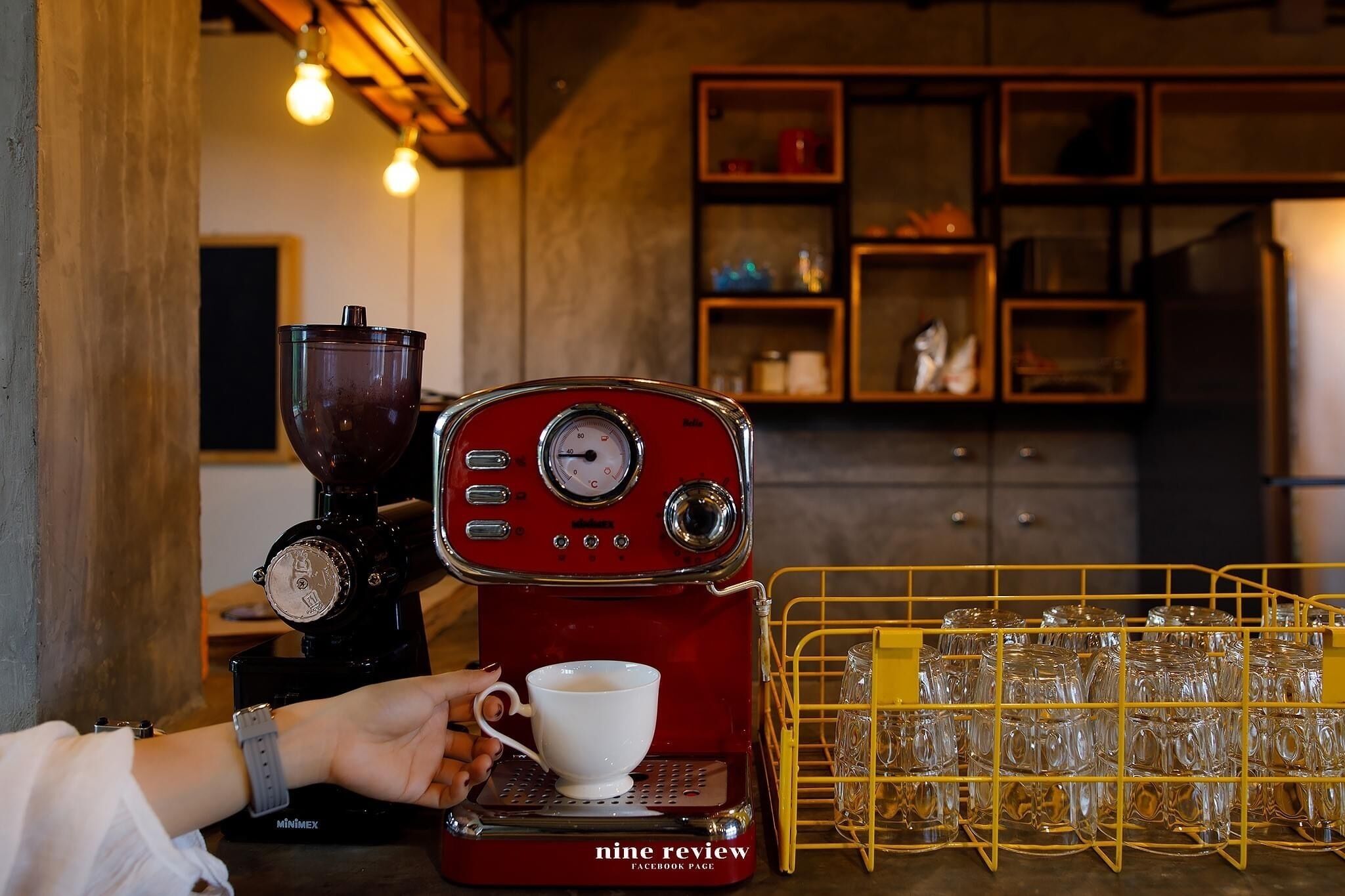 A red coffee machine with a cup of coffee being prepared. There is a yellow glass rack with multiple glasses behind it.