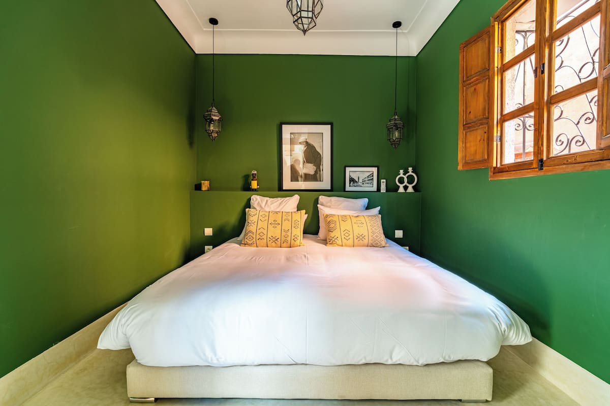 A bed with white linens and patterned pillows, a framed picture on the wall, and a window with decorative iron bars.