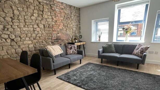 Uma sala de estar com parede de pedra, dois sofás cinzentos, uma mesa de jantar de madeira e um tapete preto.