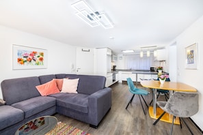 A modern living room with a grey sofa, a dining table with chairs, and a kitchen in the background.