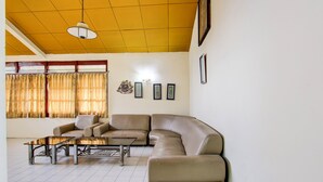 Een woonkamer met een beige hoekbank, een glazen salontafel en een kamerbreed tapijt. Drie ingelijste foto's hangen aan de muur en er is een plafondlamp.