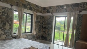 Une chambre avec un mur en pierre, un lit recouvert d’une couvre-lit à motifs, une table de chevet en bois, une chaise et un balcon donnant sur un paysage verdoyant.