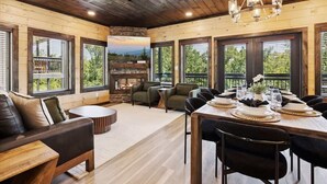 A spacious room with a wooden ceiling, large windows, a fireplace, and a dining area set for a meal. - New Modern 4BR Mtn Home w/ Indoor Pool, Rooftop Patio, Game Room, & Hot Tub (Pigeon Forge)