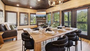 A dining area with a wooden table set for six, surrounded by black chairs, with a chandelier above and large windows with blinds. - New Modern 4BR Mtn Home w/ Indoor Pool, Rooftop Patio, Game Room, & Hot Tub (Pigeon Forge)