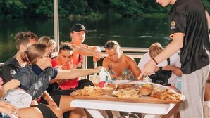 Um grupo de pessoas reunido em volta de uma mesa com comida e bebidas, sentadas ao ar livre, com um corpo de água ao fundo.