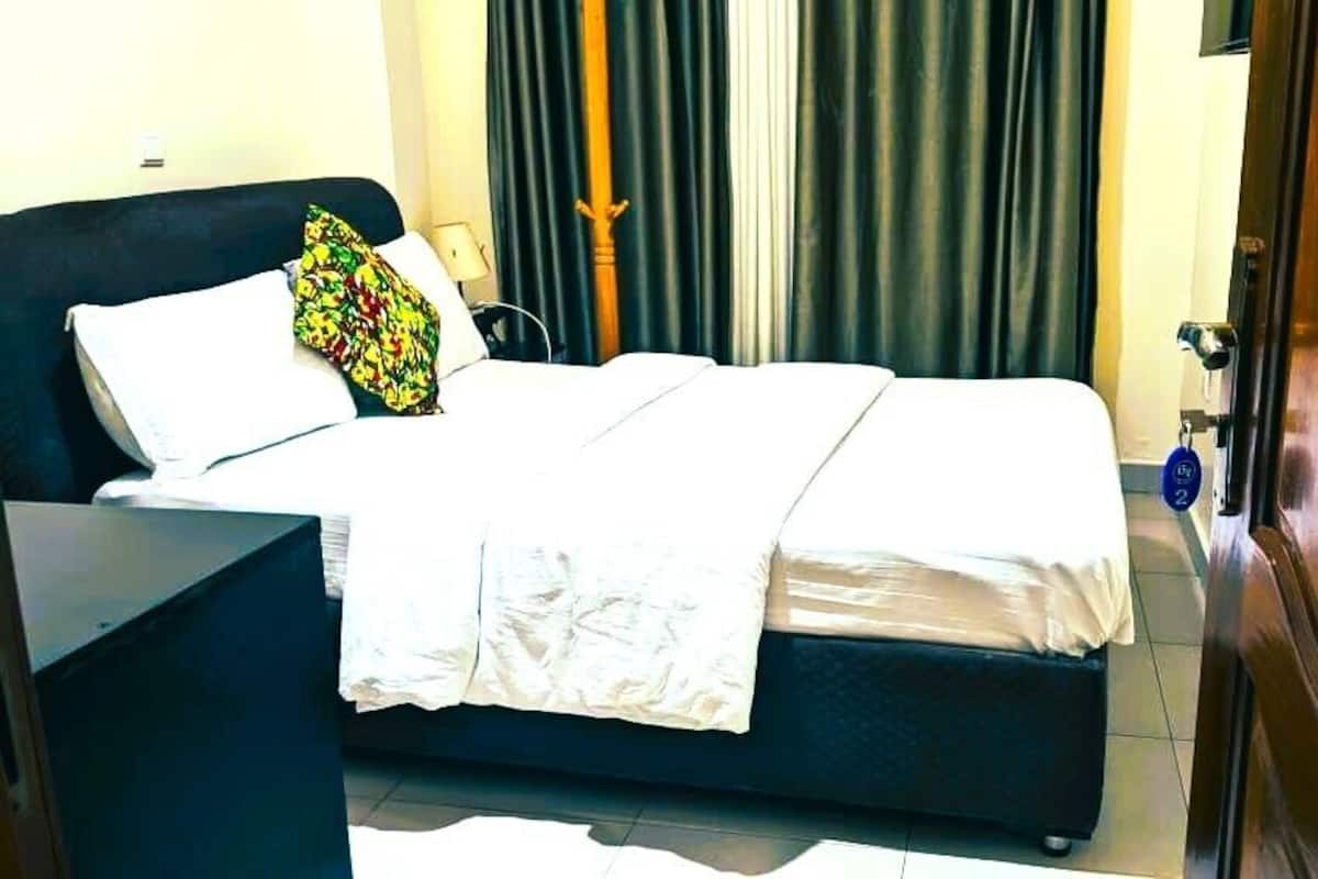 Una habitación de hotel con cama, mesita de noche, lámpara, puerta de madera y cortina.