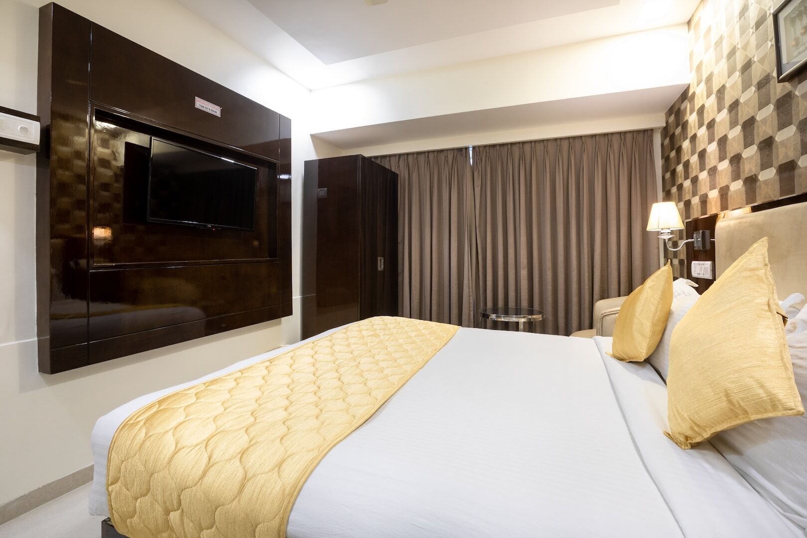 A hotel room with a large bed, a flat-screen TV mounted on the wall, a bedside table, and a lamp.