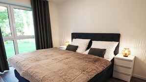 A bedroom with a large bed, two bedside tables with lamps, a window with curtains, and a view of greenery outside.