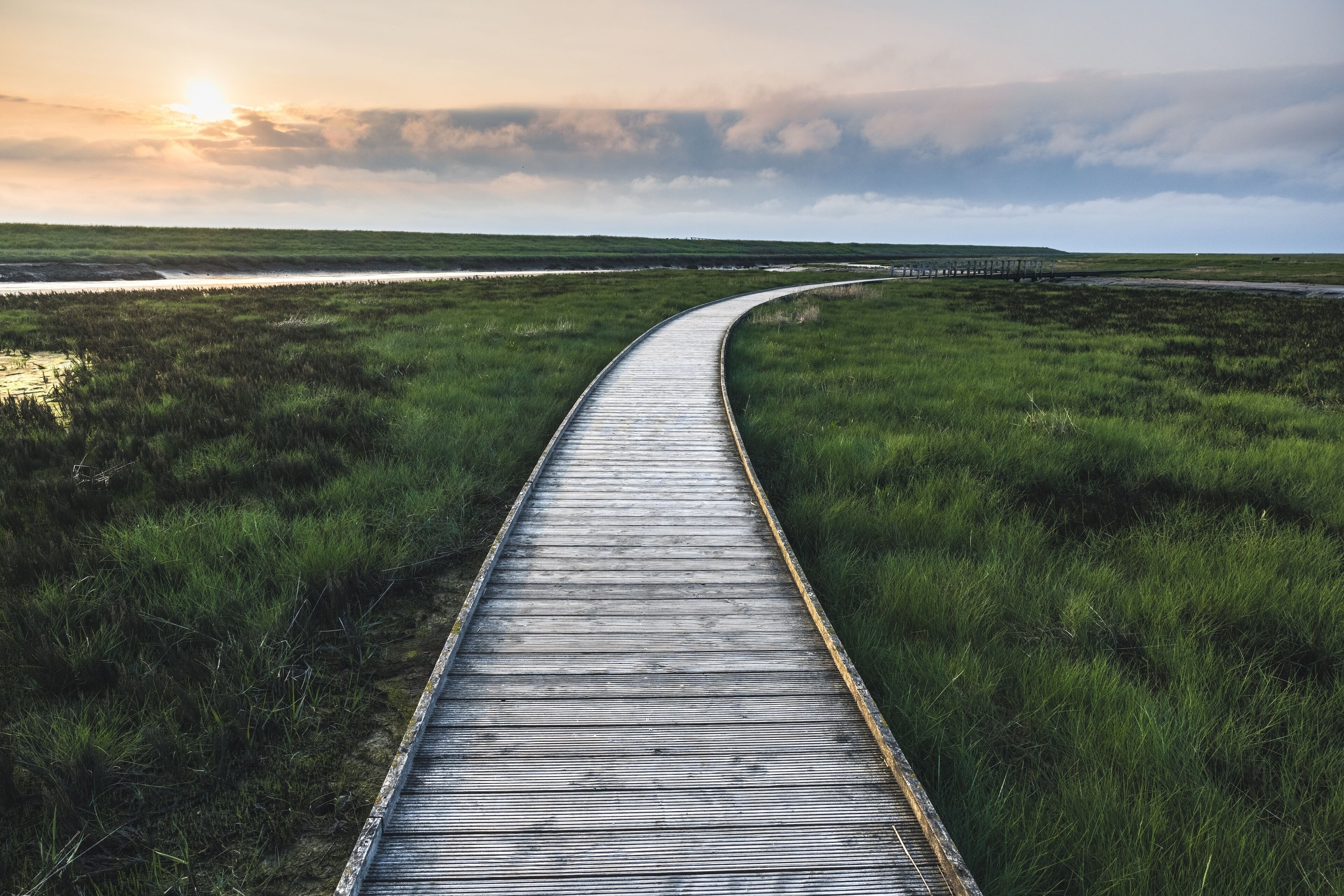 A wooden boardwalk curving through a grassy landscape with a sunset in the background.