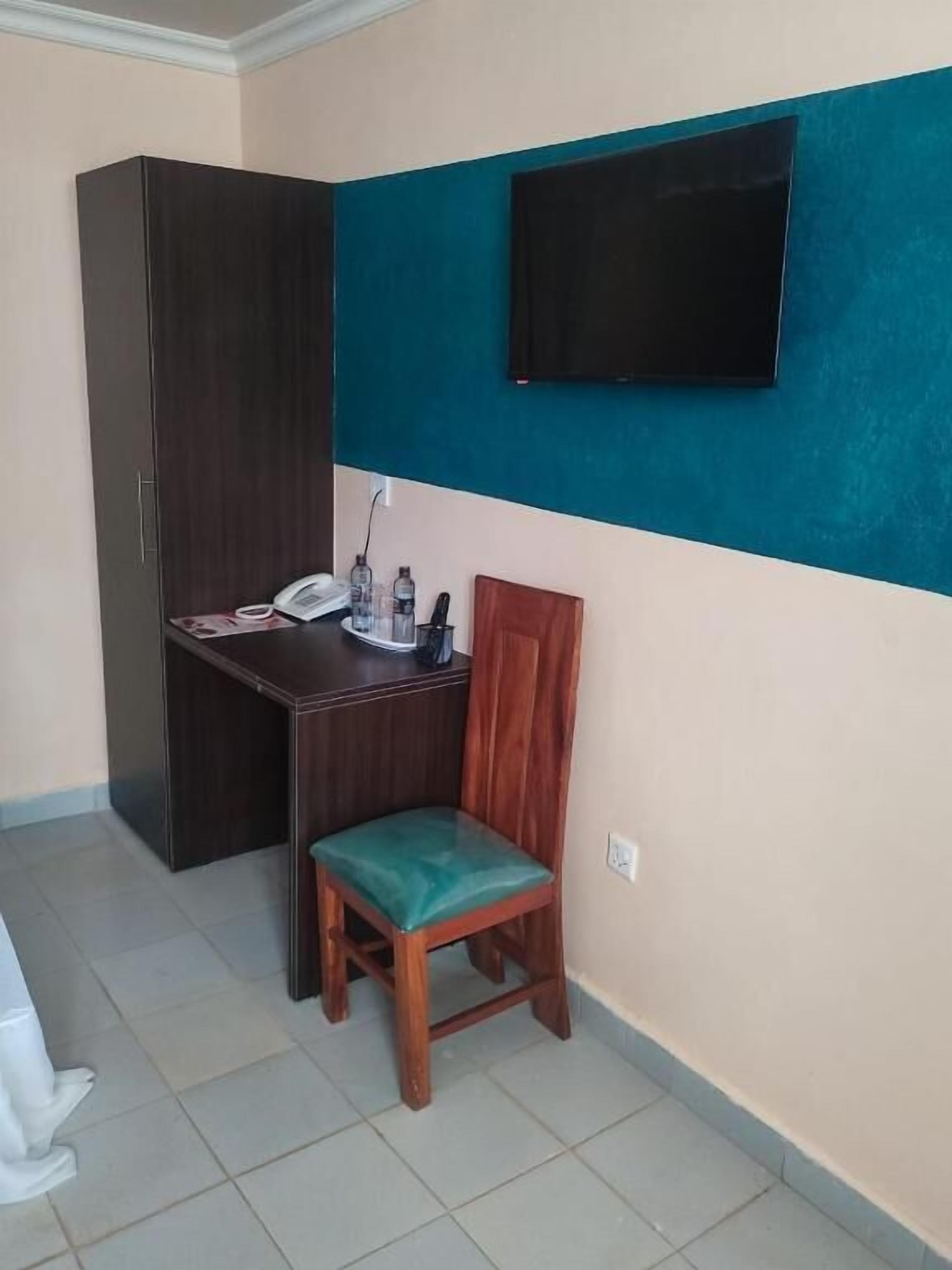 Une petite pièce avec un meuble en bois sombre, un bureau avec un téléphone, une chaise et un téléviseur fixé au mur.