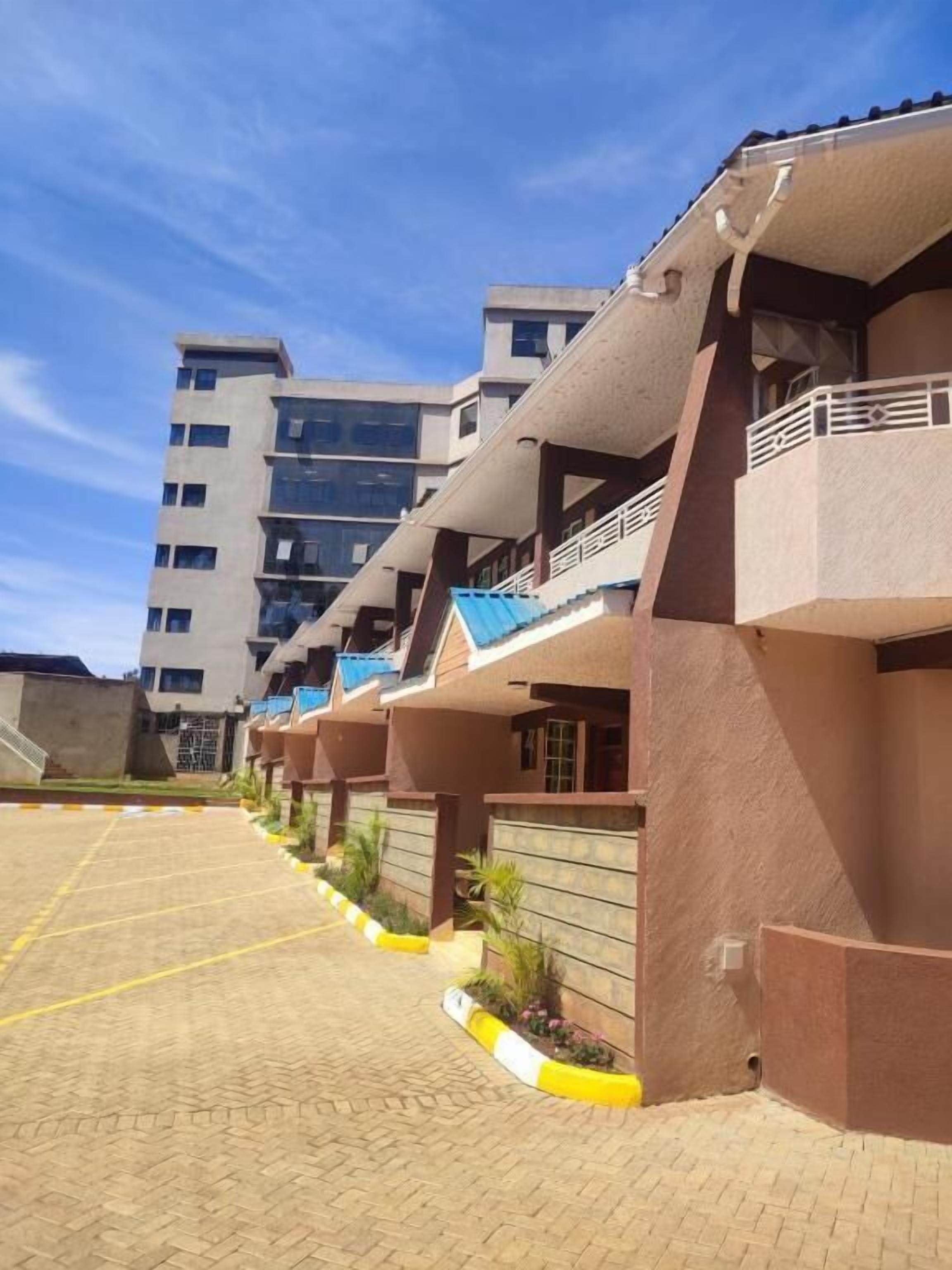 A row of modern multi-story buildings with balconies, a paved parking area, and a clear sky.