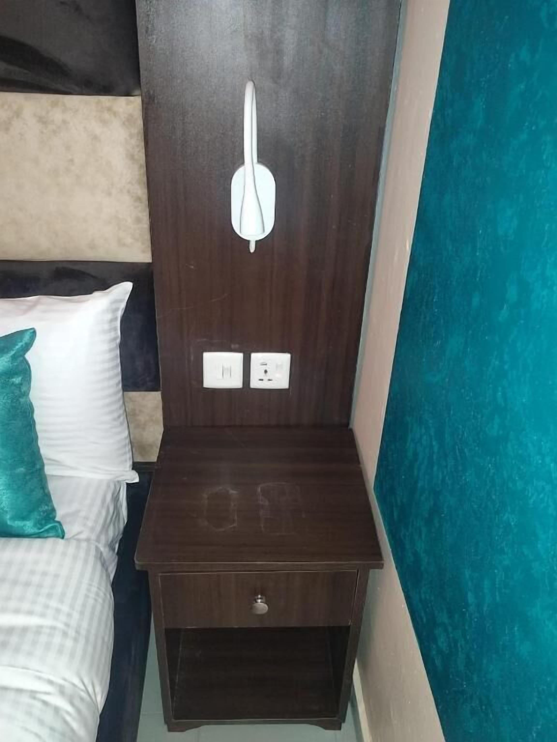 Une chambre d’hôtel avec une tête de lit en bois, un lit agrémenté d’oreillers blancs et turquoise, une petite table de chevet en bois avec un tiroir et une prise électrique au mur.
