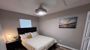 Un dormitorio con piso de madera, una cama con cubrecama a rayas, una mesita de noche con lámpara, una ventana con persianas y un cuadro en la pared.