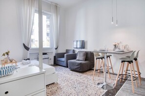 Une pièce moderne et bien éclairée, avec un canapé blanc, une table à manger et des tabourets de bar, un téléviseur à écran plat fixé au mur et une fenêtre agrémentée de rideaux blancs.