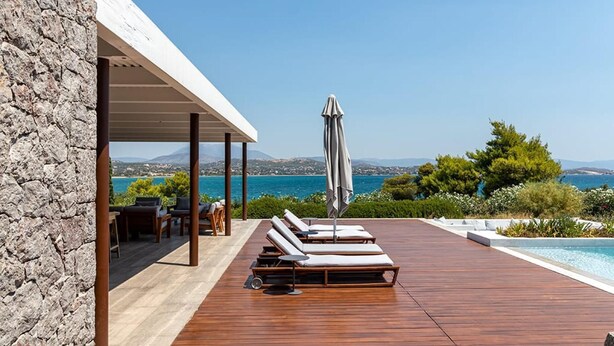 A wooden deck with lounge chairs and an umbrella, overlooking a pool and a scenic view of the sea and hills.