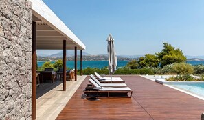 Une terrasse en bois avec des chaises longues et un parasol, offrant une vue sur une piscine et un paysage pittoresque de la mer et des collines.