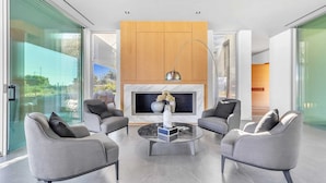 A modern living room with a fireplace, two grey armchairs, a coffee table, and a large window offering a view of the outdoors.