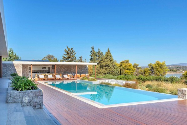 A modern outdoor pool area with a stone deck, lounge chairs, and a view of trees and a lake.