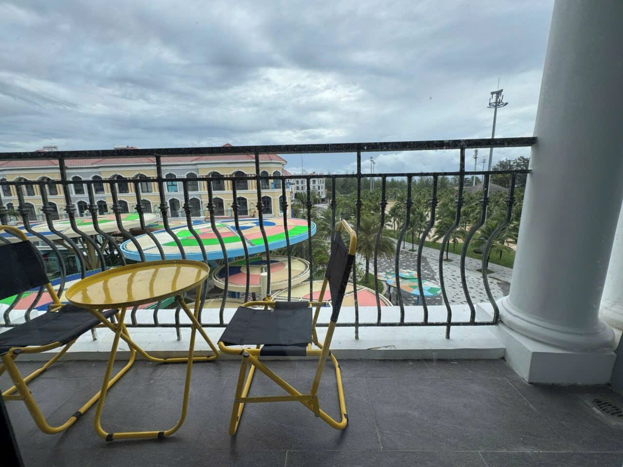 A balcony with yellow chairs and a table, overlooking a colorful playground and buildings.