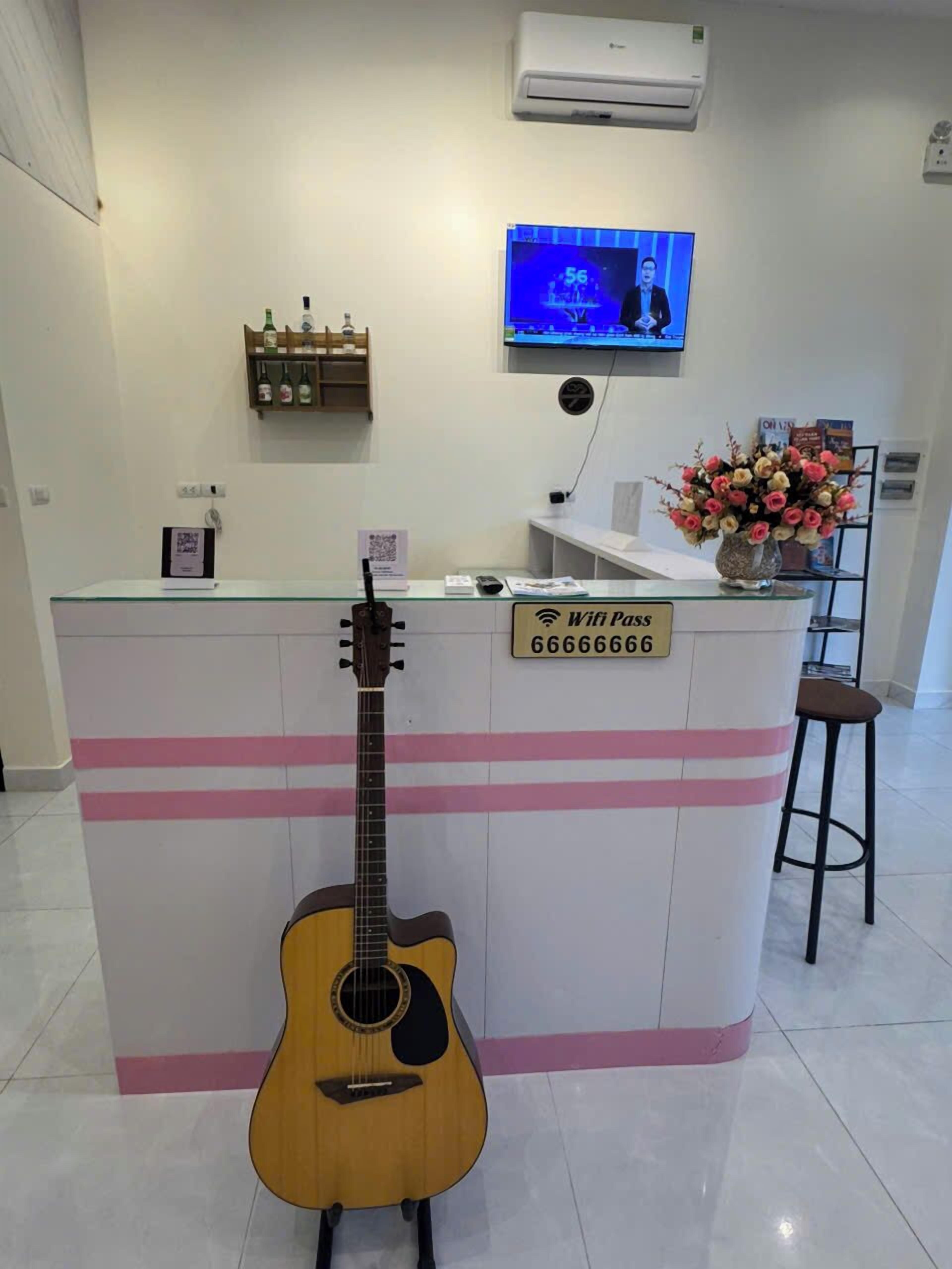 A reception area with a guitar, a TV displaying a news program, a Wifi pass sign, and a flower vase.