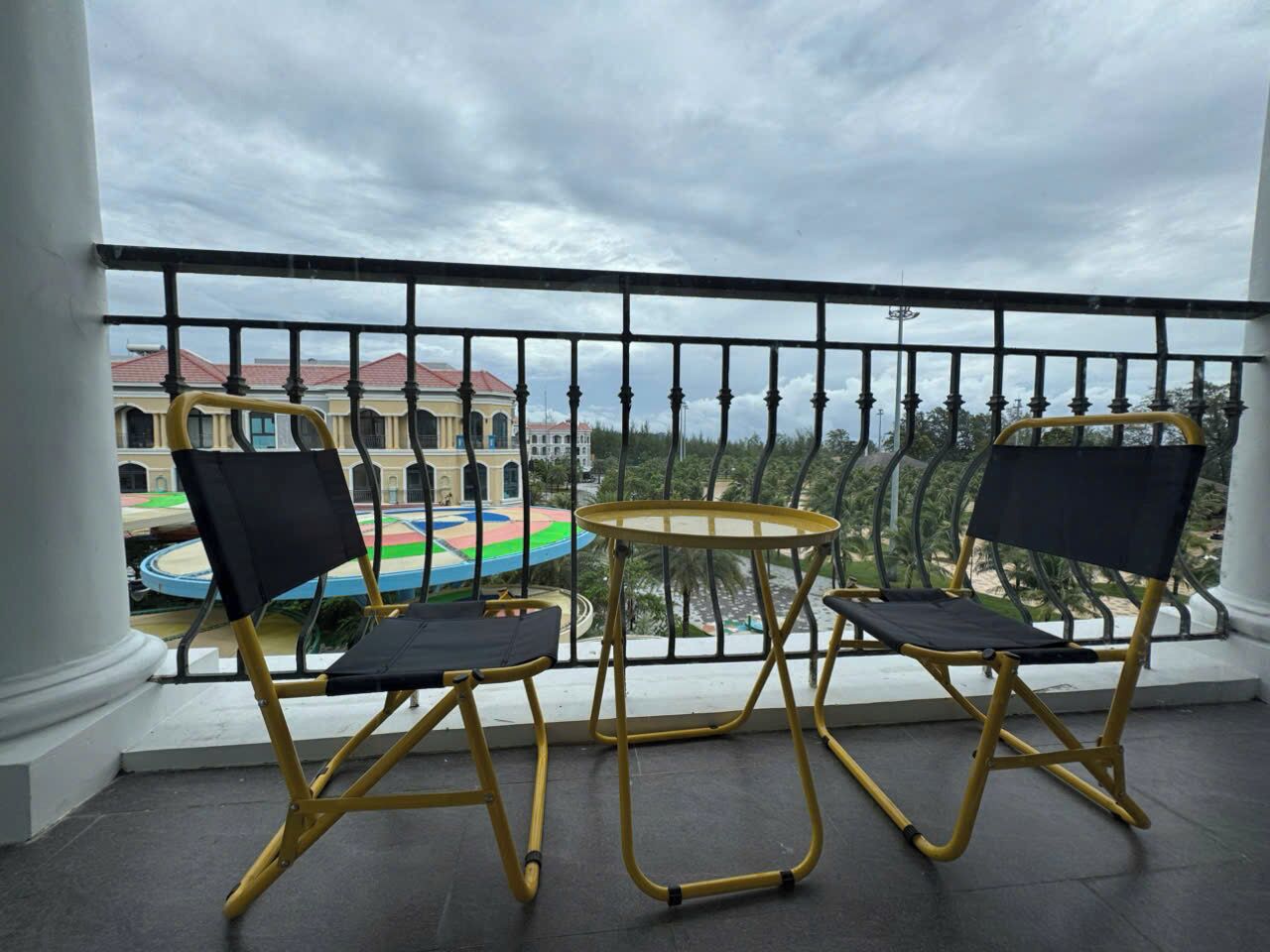 Un balcón con dos sillas y una mesita, con vistas a un parque infantil colorido y edificios.