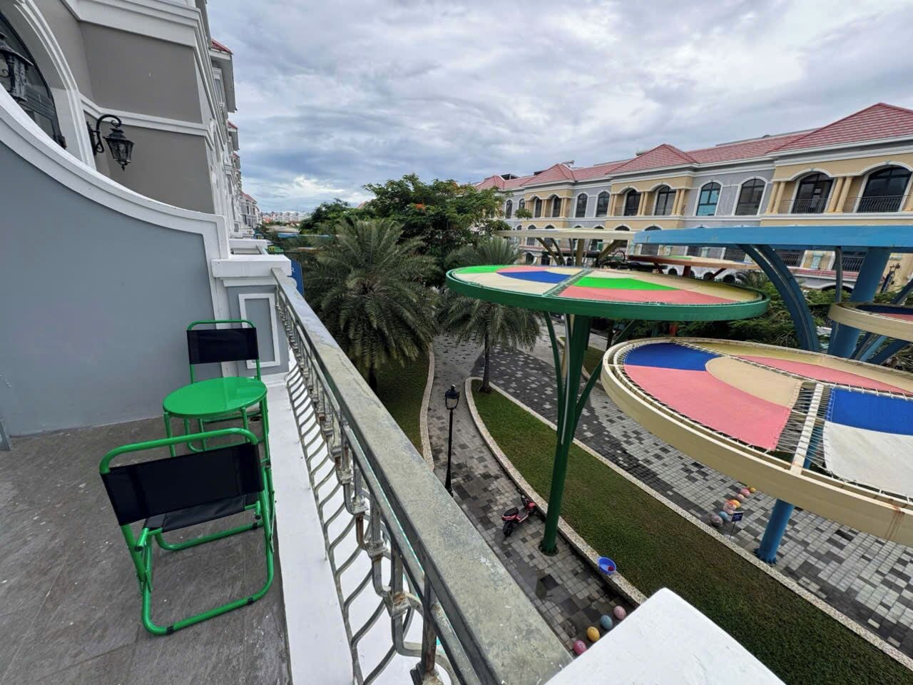 A balcony with green chairs, a small table, and a view of a playground with a colorful play structure.