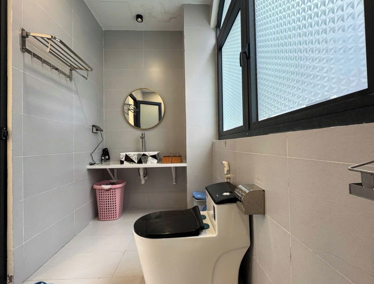 Un baño moderno con inodoro, lavamanos y espejo. Cuenta con una ventana con persianas, un perchero y una cesta de ropa.