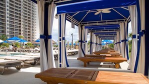 Een zwembadterras met houten lounge tafels en parasols, met hoogbouw op de achtergrond en een heldere lucht.
