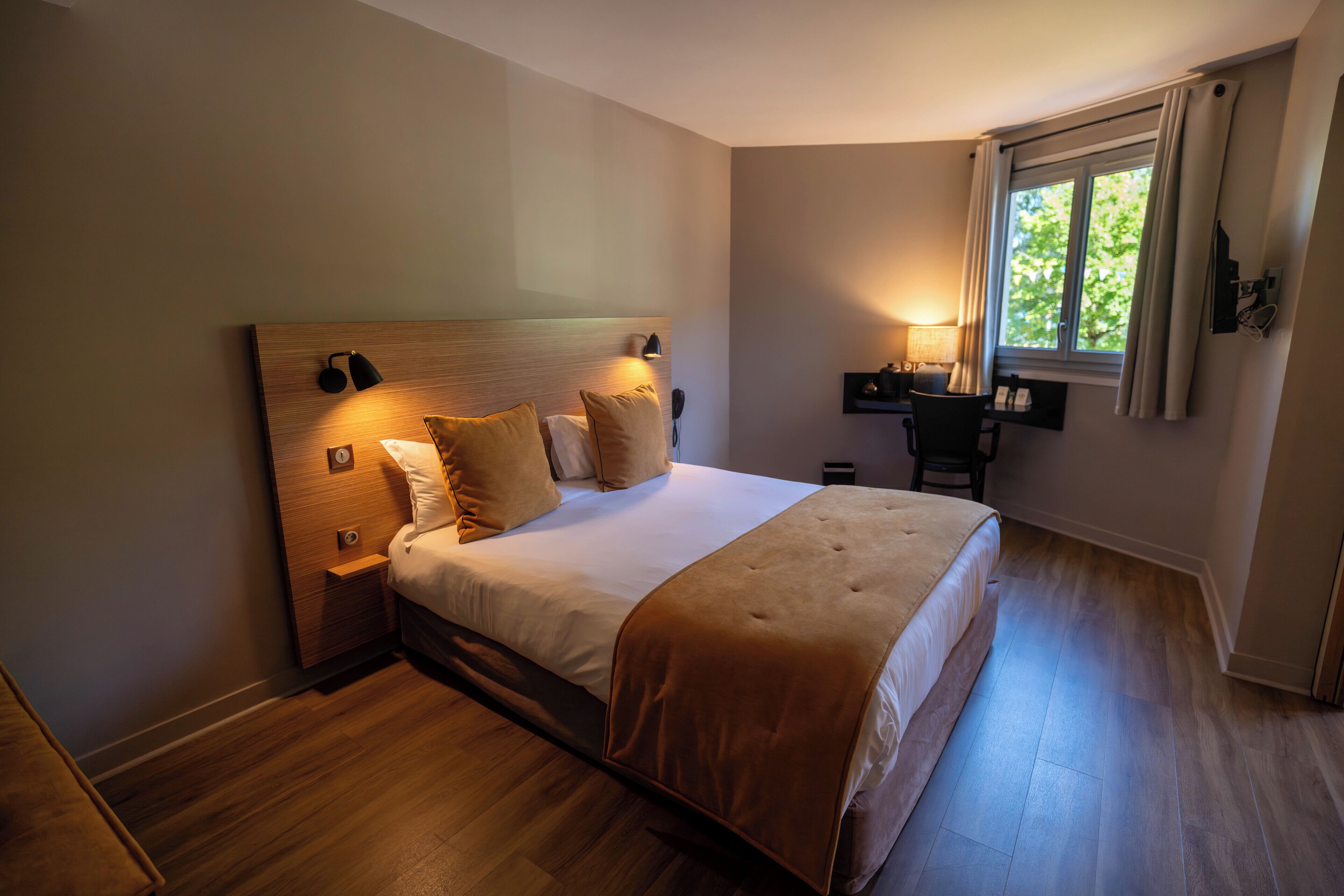A modern bedroom with a wooden headboard, a large bed with a beige blanket, a bedside table with a lamp, and a window with curtains.