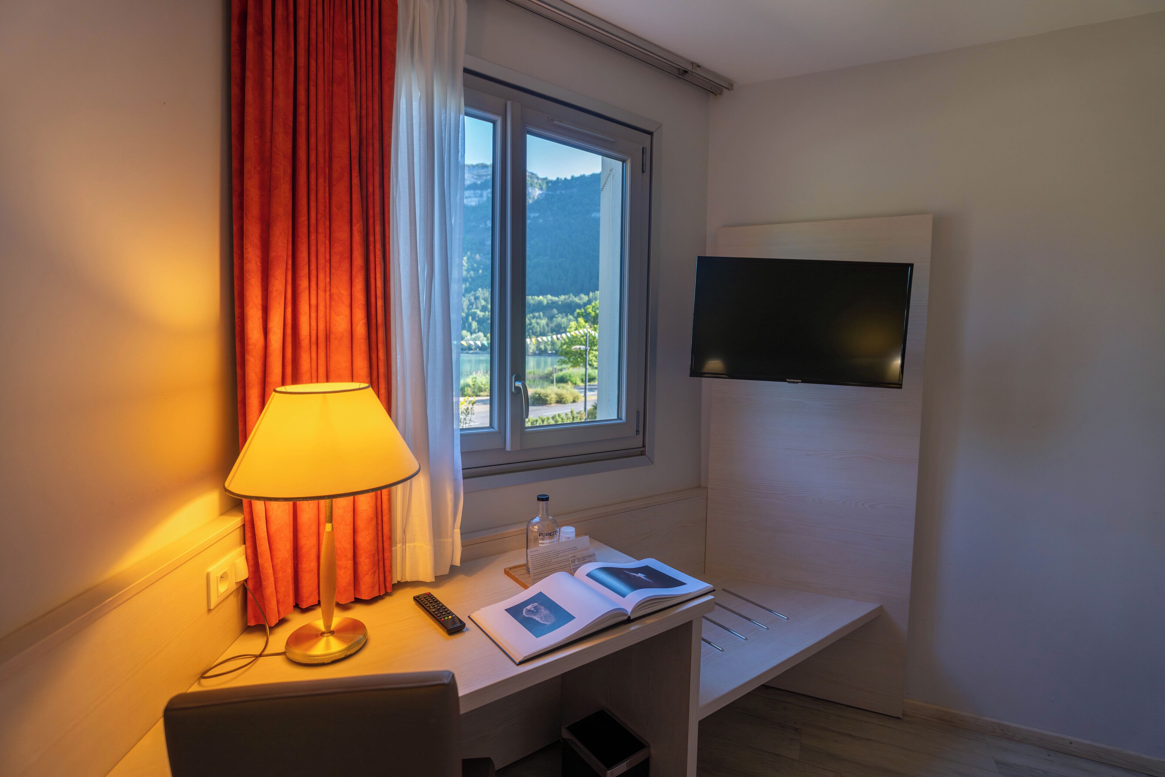 Ein Hotelzimmer mit Schreibtisch, Lampe, Fernbedienung, geöffnetem Buch, Flasche und Fernseher auf einem Tisch.