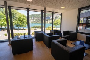 Un área de estar moderna con sofás de cuero negro y mesas, con amplios ventanales que ofrecen vistas a un lago y montañas.