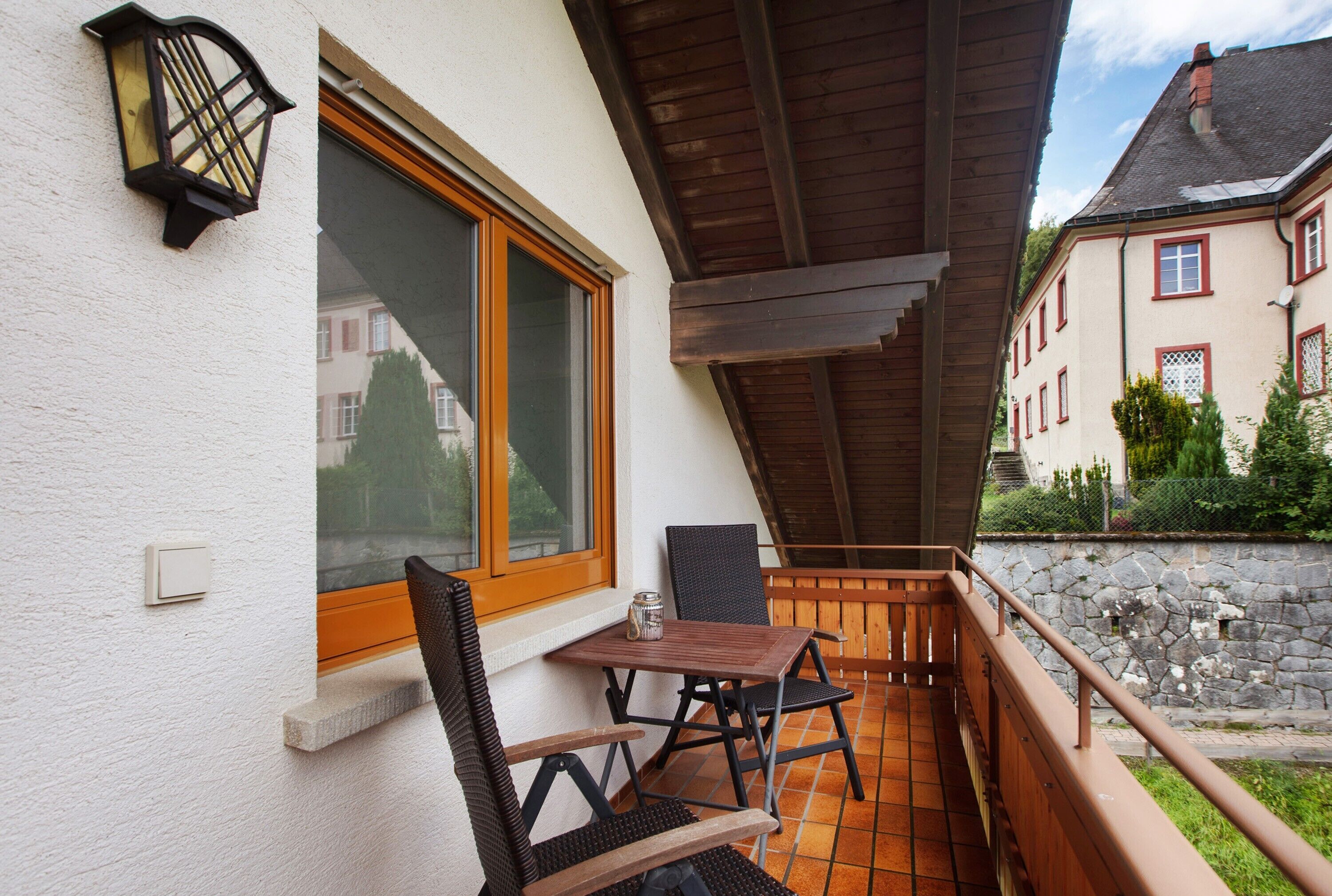 A balcony with a table and chairs, a window, and a stone wall in the background.
