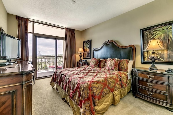 A spacious bedroom with a large bed, a dresser, a television, and a balcony with a view.