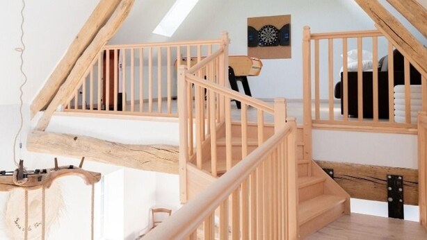 A wooden staircase with a simple railing, leading to a loft bedroom with a bed and a dartboard on the wall.
