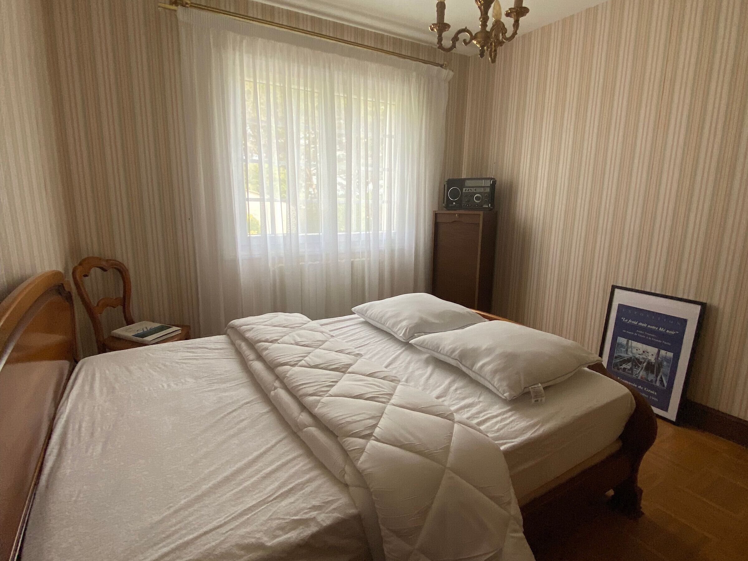 A neatly made bed with a quilted cover, a wooden nightstand with a book, a radio on a cabinet, and a framed picture on the wall.